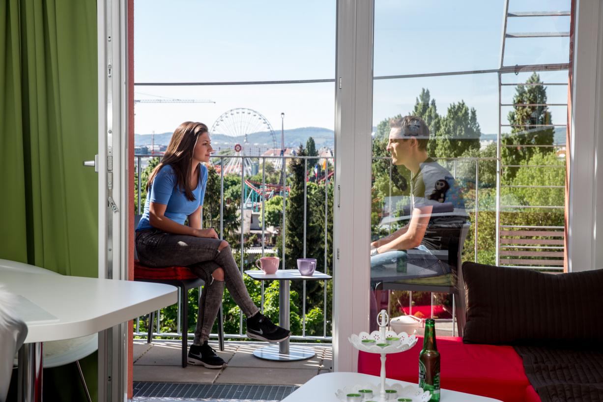 Two students are sitting on a sunny balcony. In the background we can see a giant wheel in the Prater.